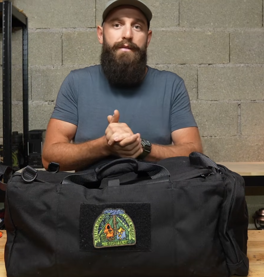 A man wearing a blue-gray t-shirt and a brown cap stands behind a Savotta Keikka 50L duffel bag. The bag features a Cult of Savotta patch on the front.