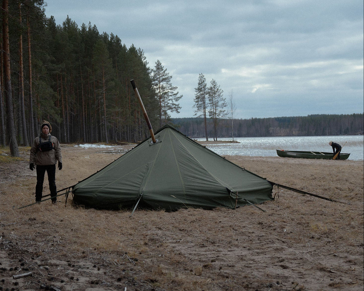 Person standing next to Savotta Hawu 8 tent on a lakeside beach with trees and water in the background.