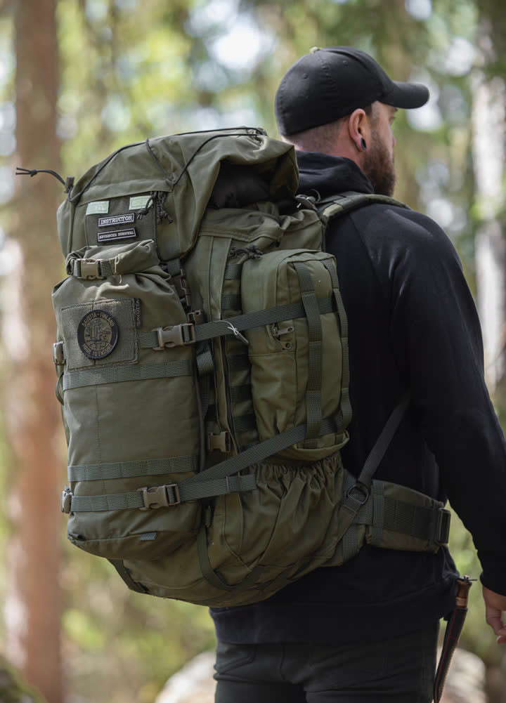 Person wearing a green Kantamus 60L backpack with Hatka 12L attached to the front and side 4L pouches in a forest setting
