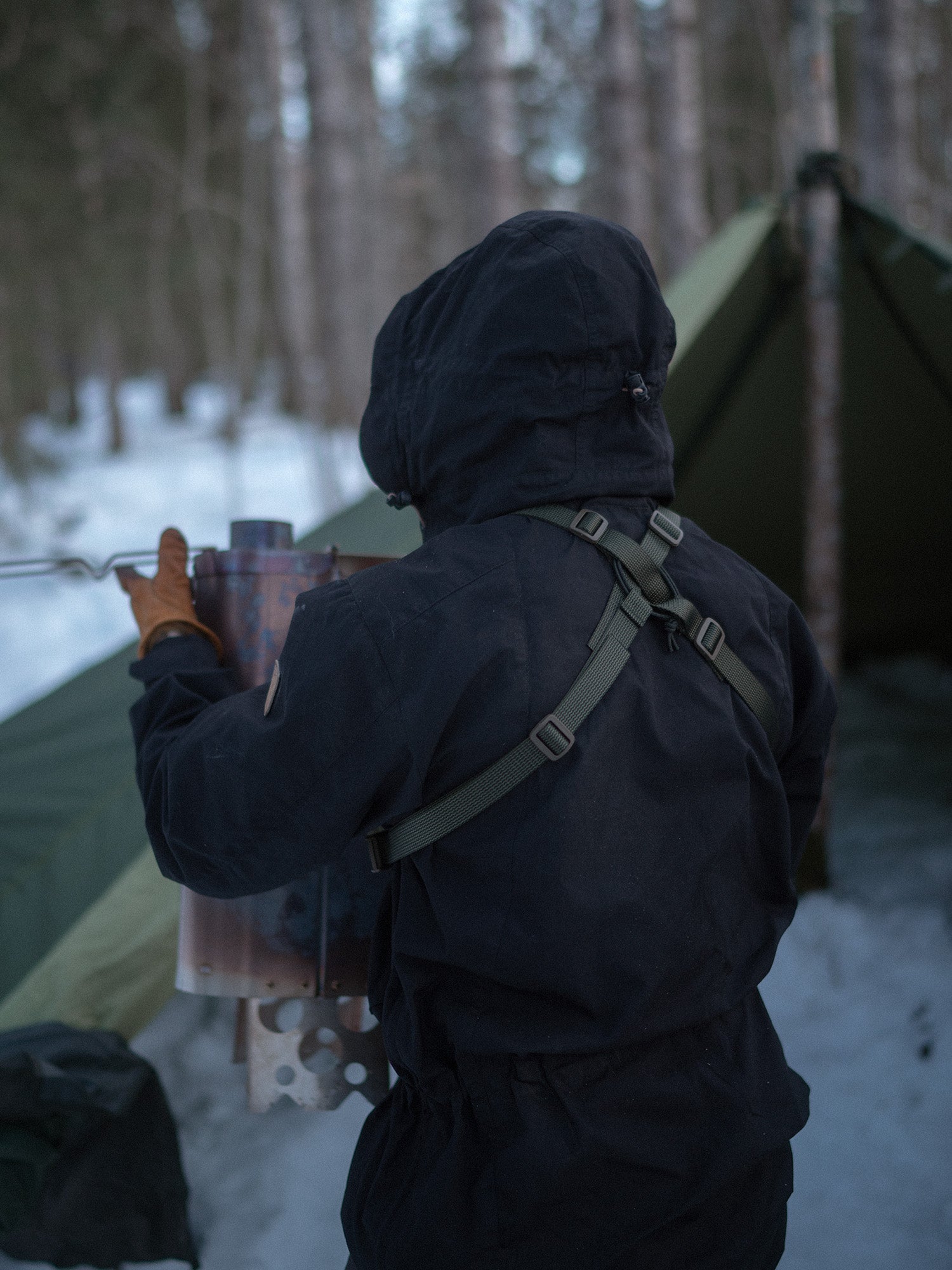 Person in dark winter clothing holding a Savotta tent stove in a snowy forest setting