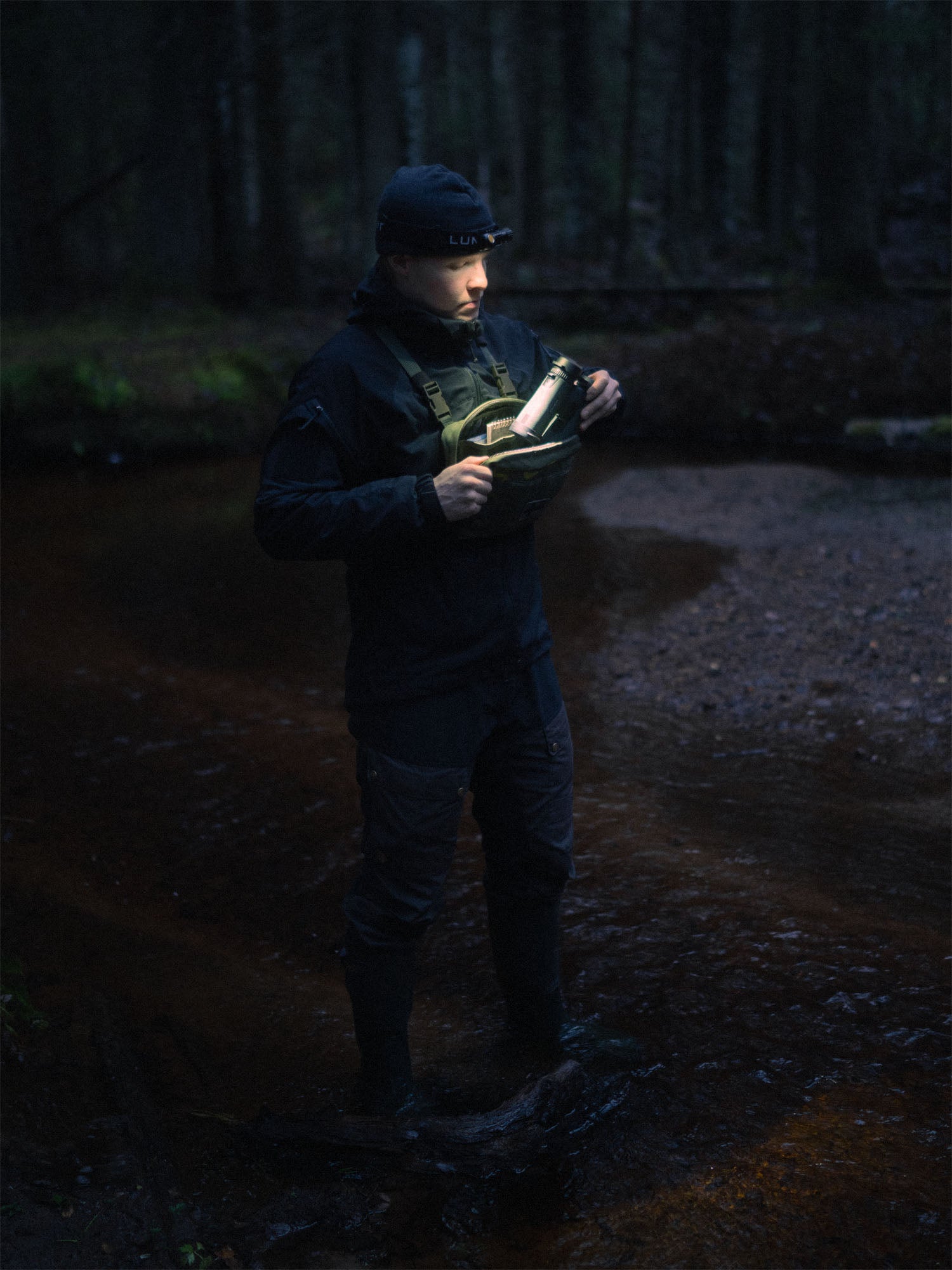 Person in dark clothing holding binoculars in a forest at night and wearing a Savotta Rintamus chest pack.