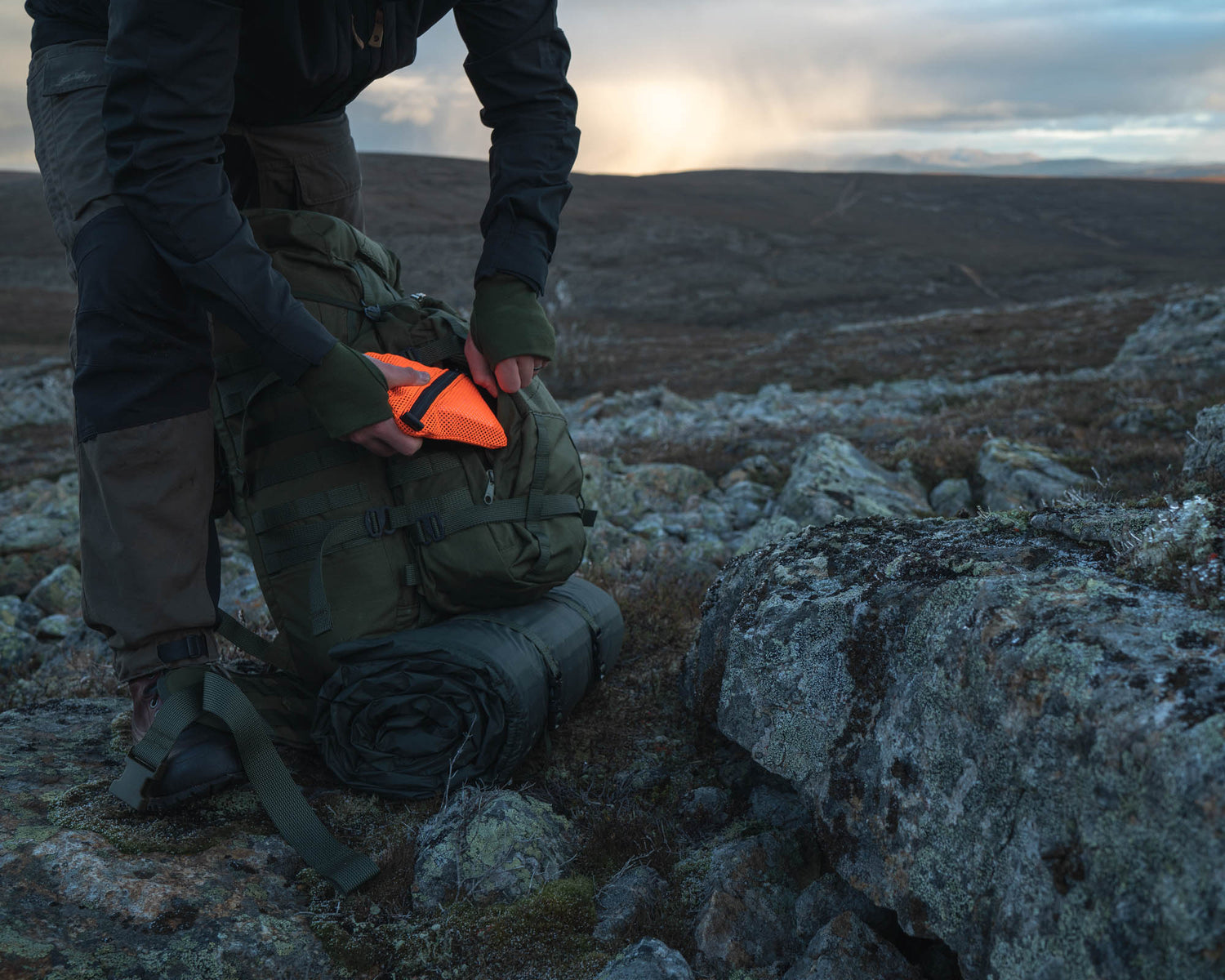 Person packing a Jääkäri backpack with the Savotta Trinket pouch in orange, on a rocky landscape with a sunset in the background
