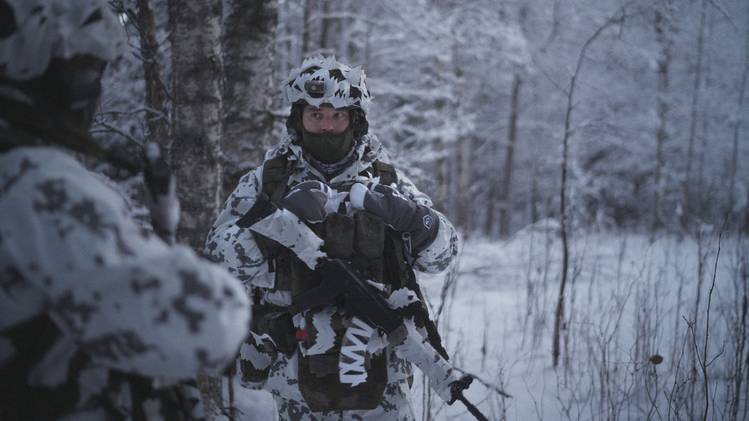 Person in Finnish M05 snow camouflage gear with Savotta helmet camo scrim in white on helmet and Savotta Varjelus Plate carrier in M05 Woodland in a snowy forest