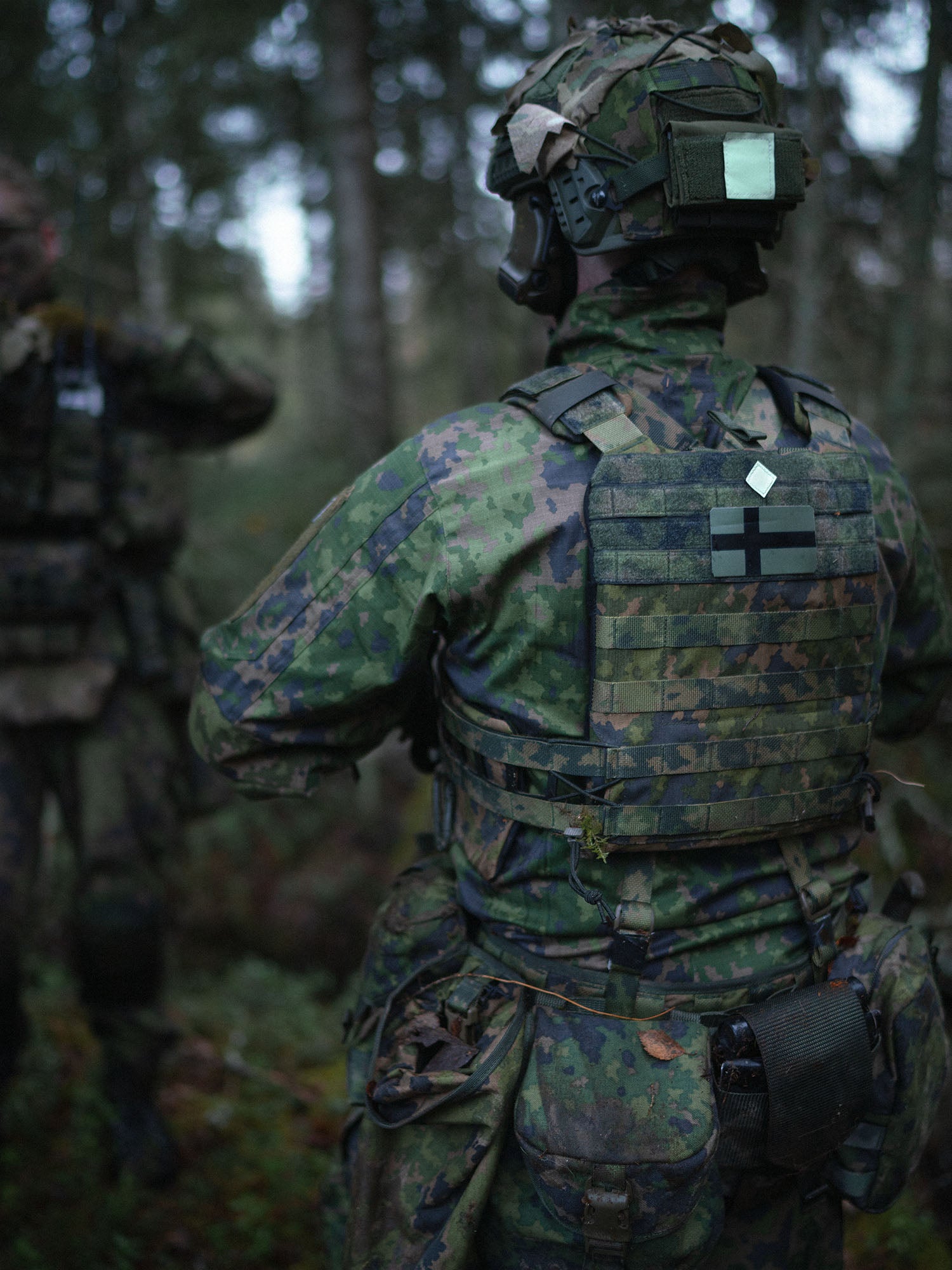 Person in camouflage military uniform with a Savotta Varjelus plate carrier on top, in a forest setting