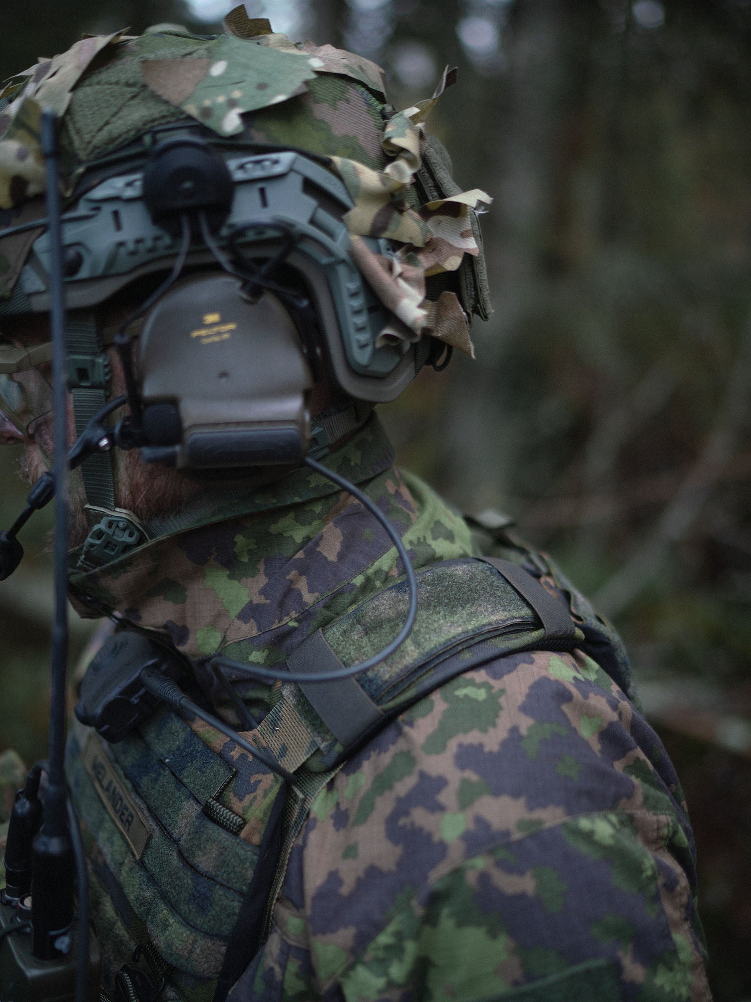 Person wearing full camouflage military gear and Varjelus plate carrier with shoulder pads, in a forest setting