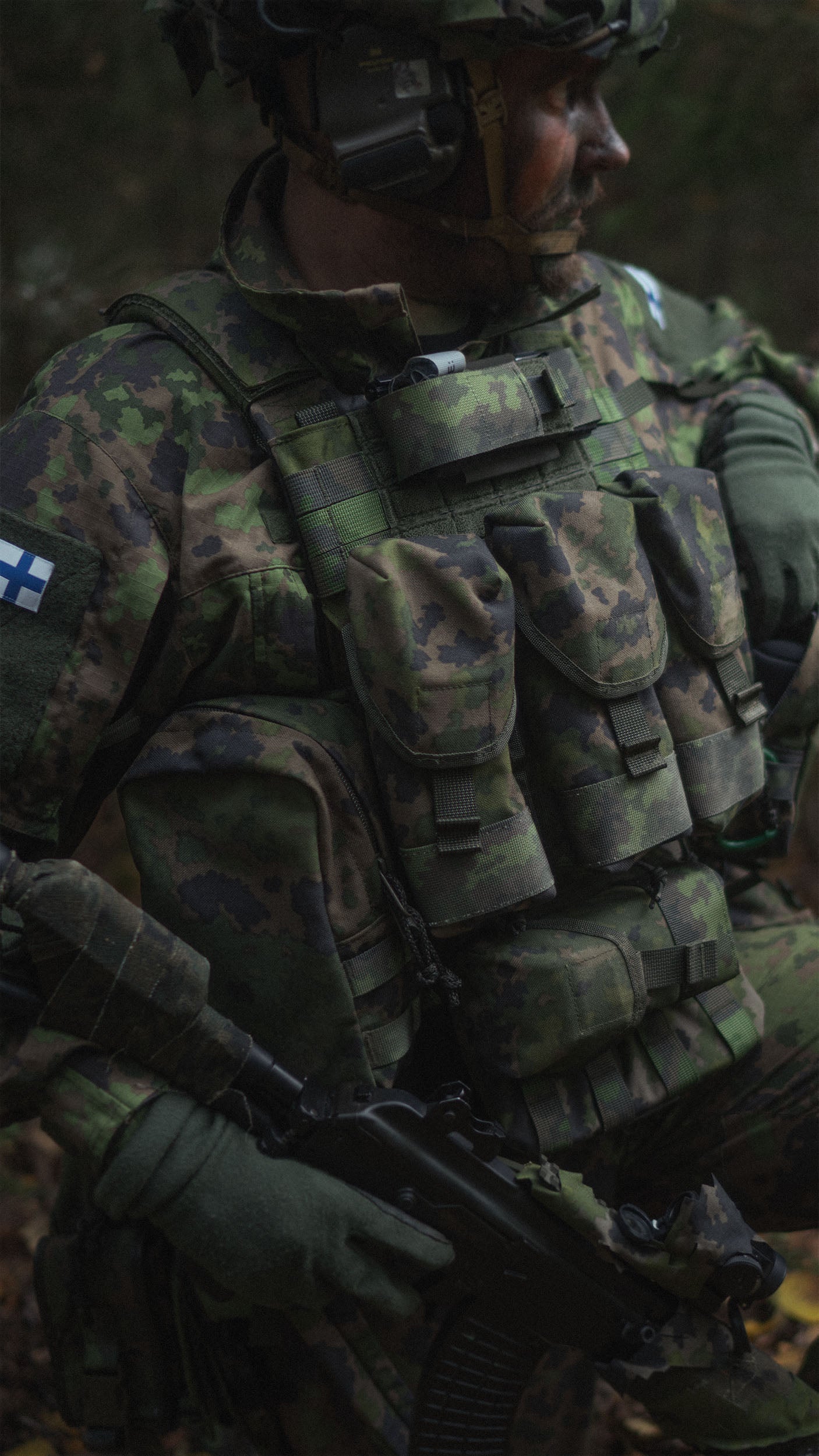Person in military camouflage gear with a Savotta Varjelus plate carrier. There are multiple pouches attached to the plate carrier. A dark forest background
