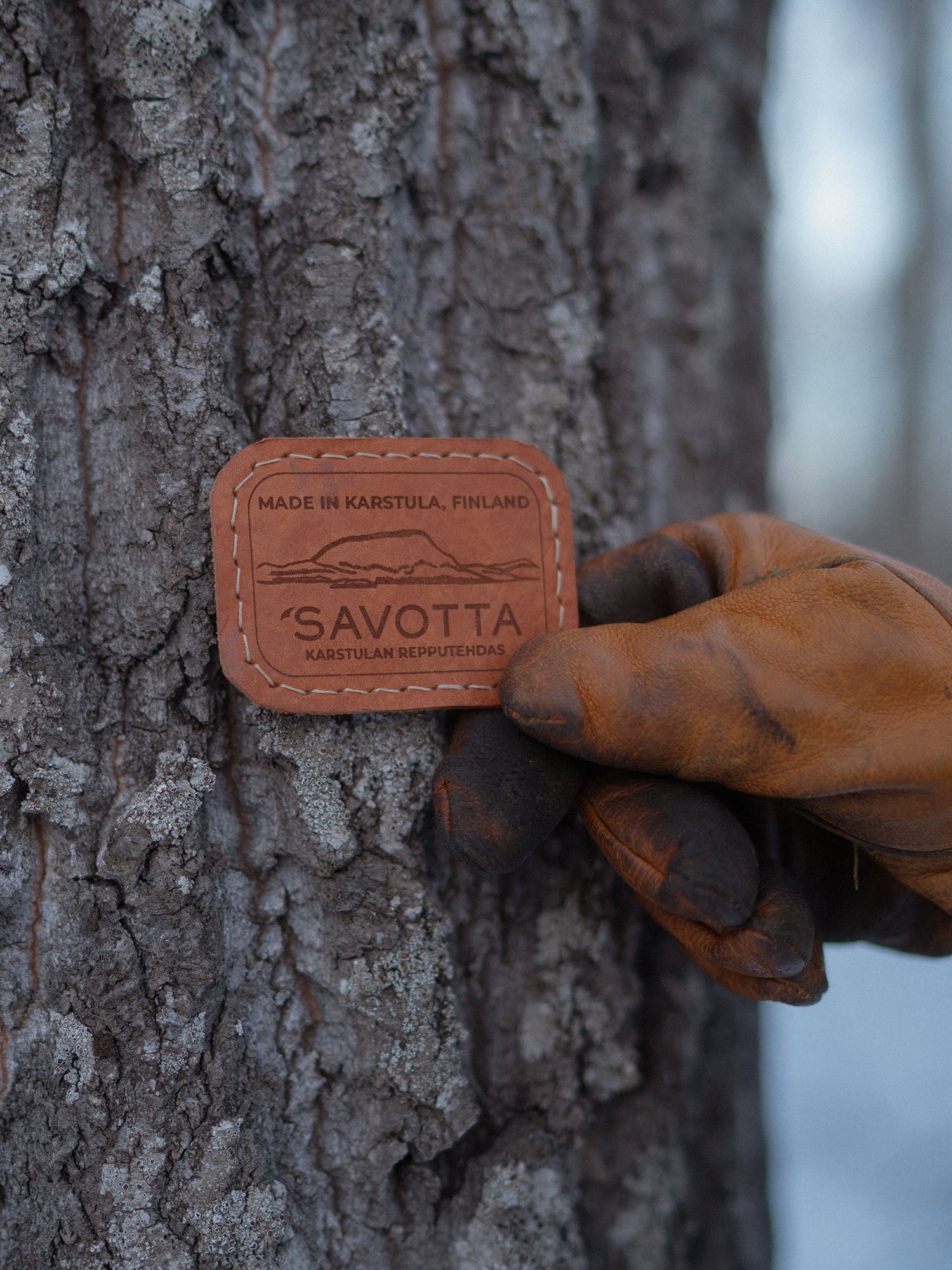 Hand wearing a brown glove holding a leather patch with 'Savotta' branding against a tree.
