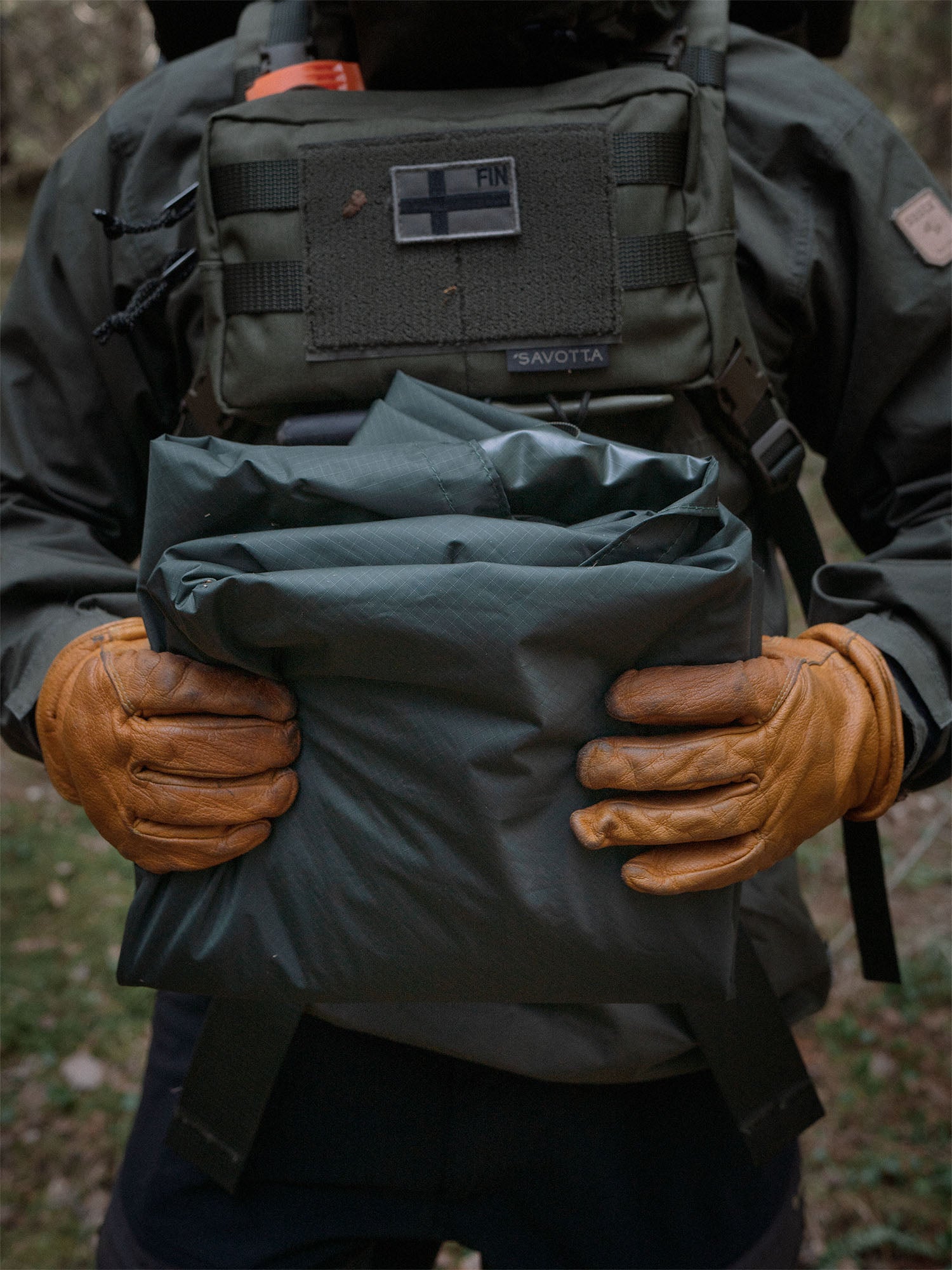Person wearing a Savotta Rintamus chest pack and holding a packed Savotta Laavu tarp in an outdoor setting