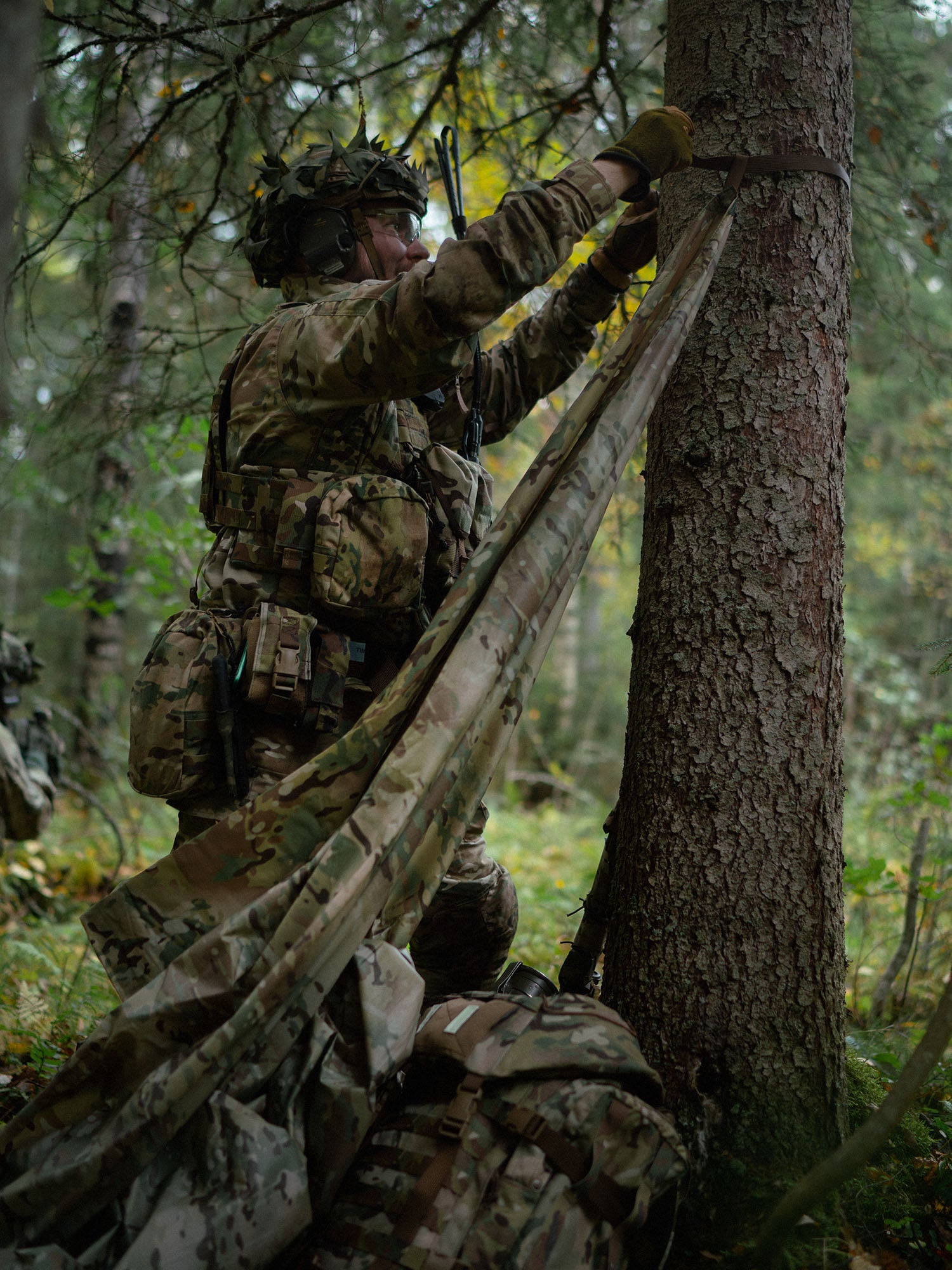 Person in military camouflage gear attaching a Savotta Multicam Laavu tarp to a tree in a forest setting