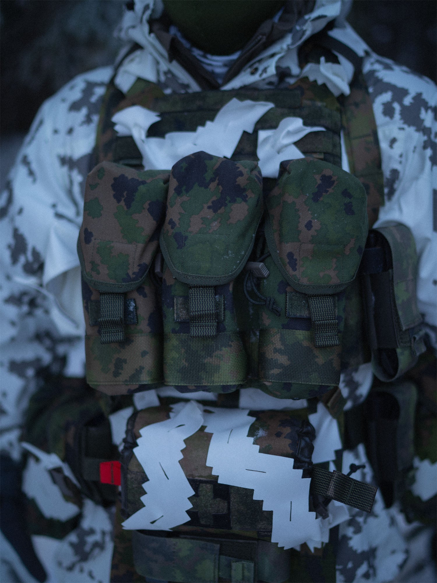 Person wearing camouflage military gear and a Savotta Varjelus plate carrier with Varjelus 3x2 AR15-AK placard attached, snowy setting