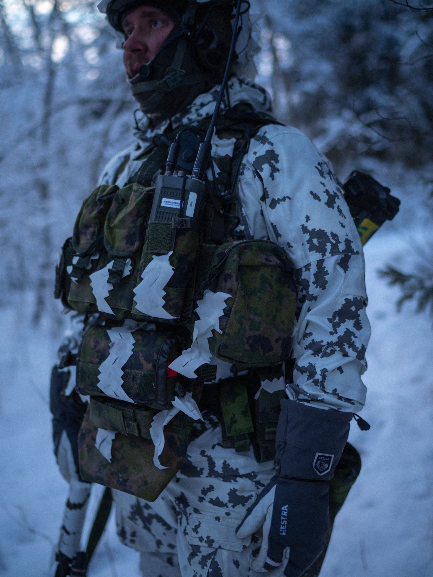 Person in camouflage gear and a Savotta Varjelus plate carrier with a Varjelus 3x2 AR15-AK placard and Varjelus groin flap attached, standing in a snowy forest