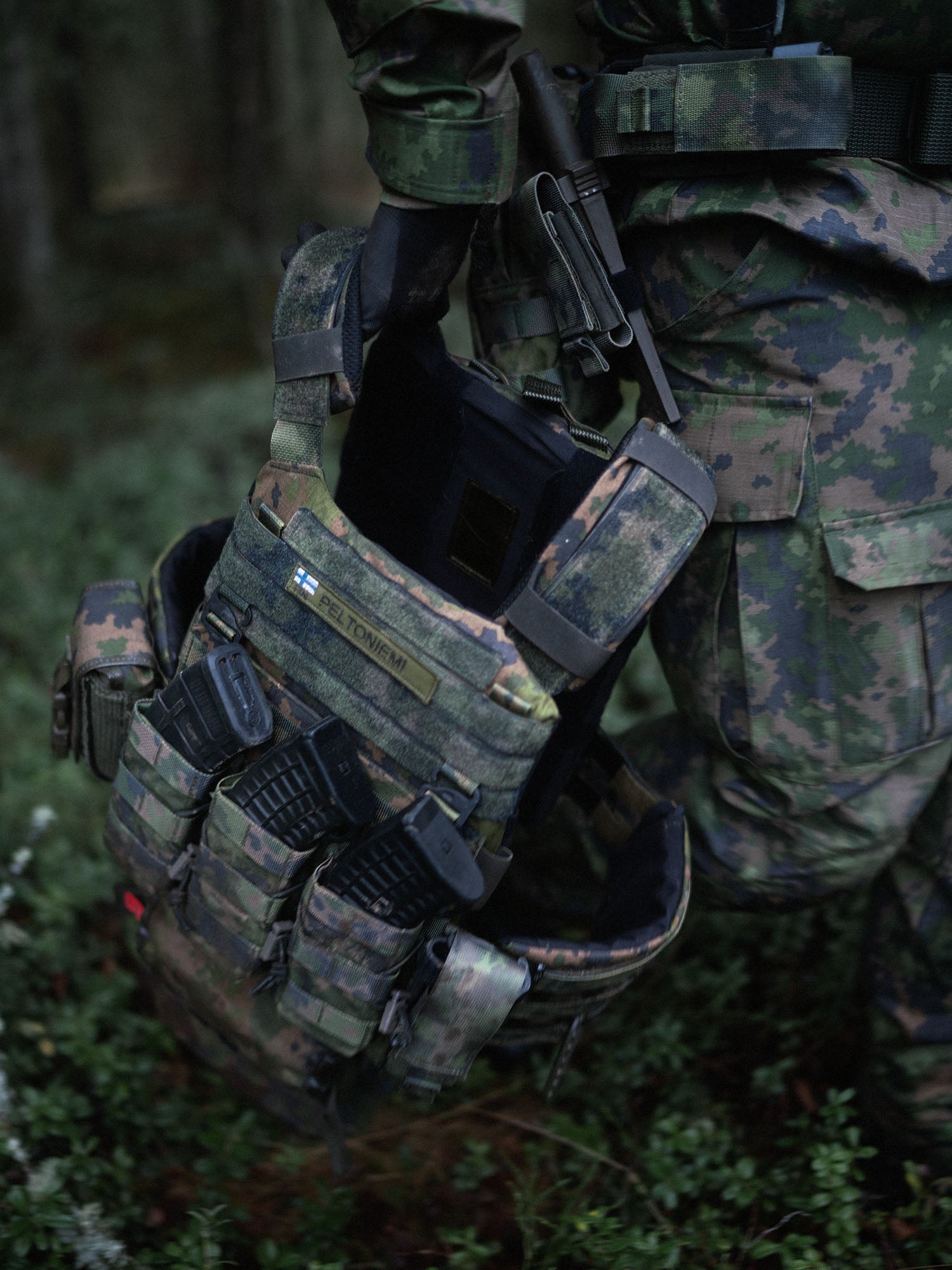 Savotta Varjelus plate carrier with mag pouches and a Varjelus groin flap attached, being carried by a person in camouflage gear on a forest background