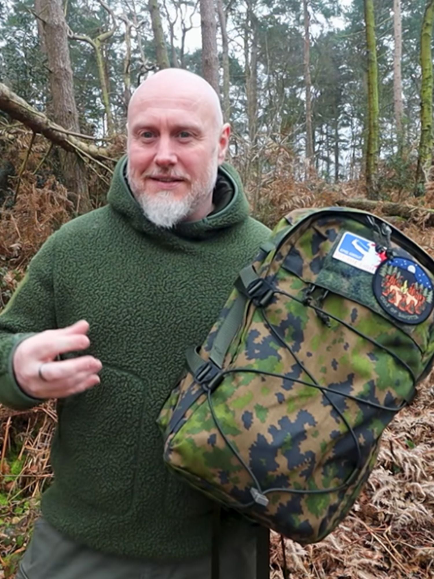 Man in a forest wearing a green shirt holding a Niukka 20L camo backpack