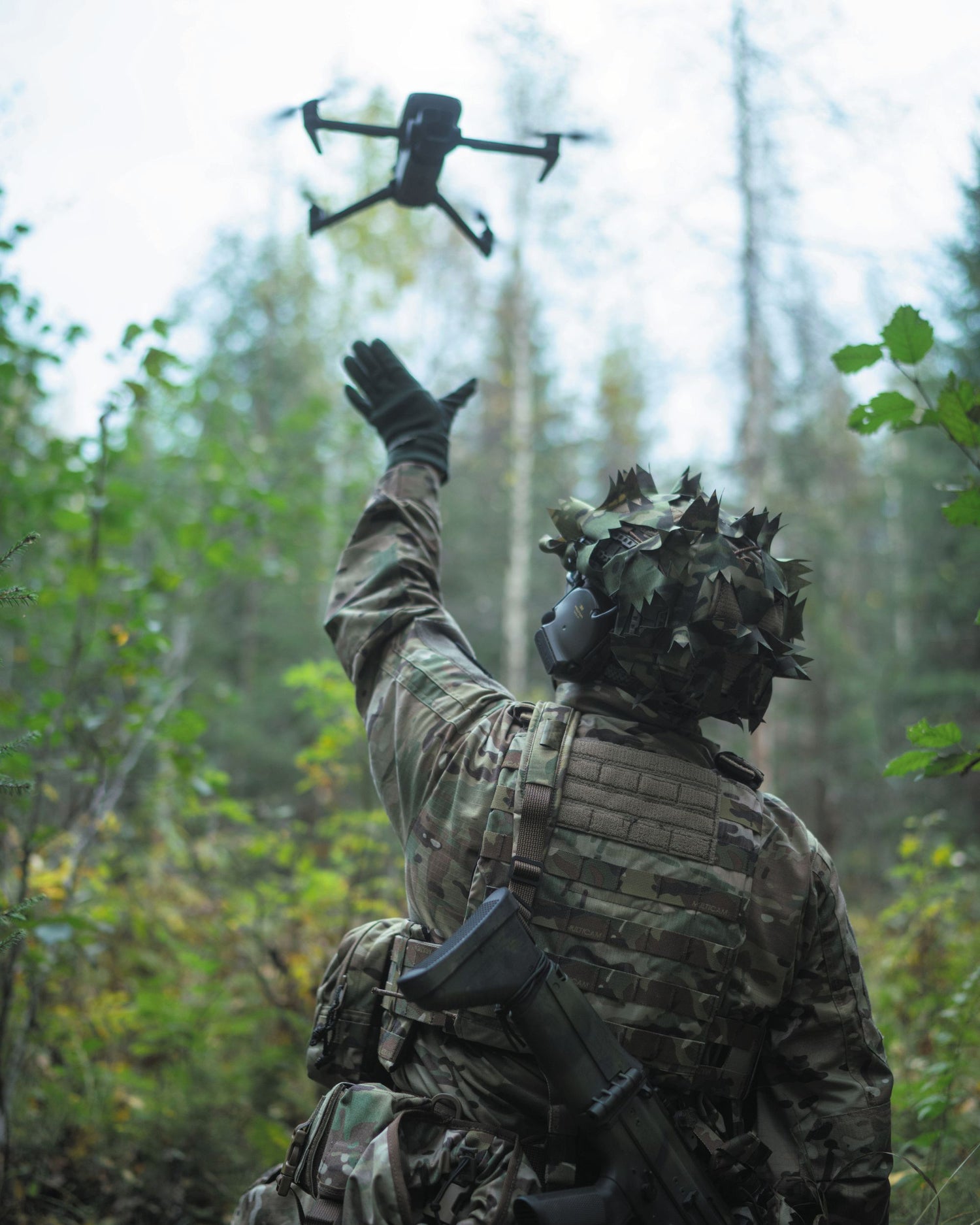 Person in military gear flying a drone in a forest setting