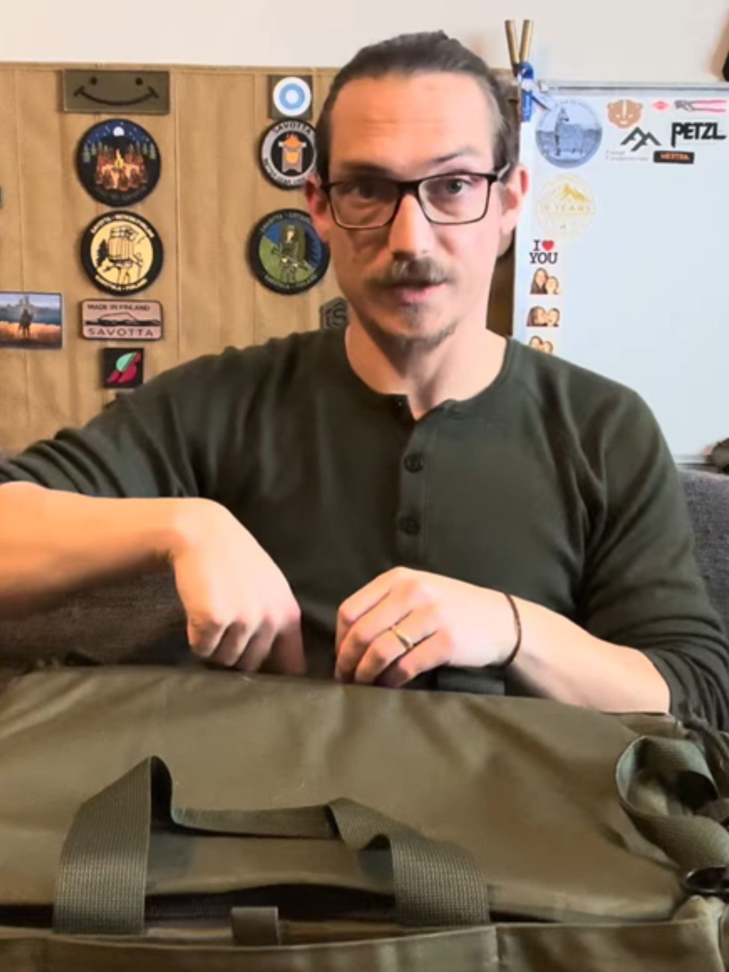 Man sitting with a green Savotta Keikka 50L duffel bag in front of him, wearing glasses and a dark shirt.