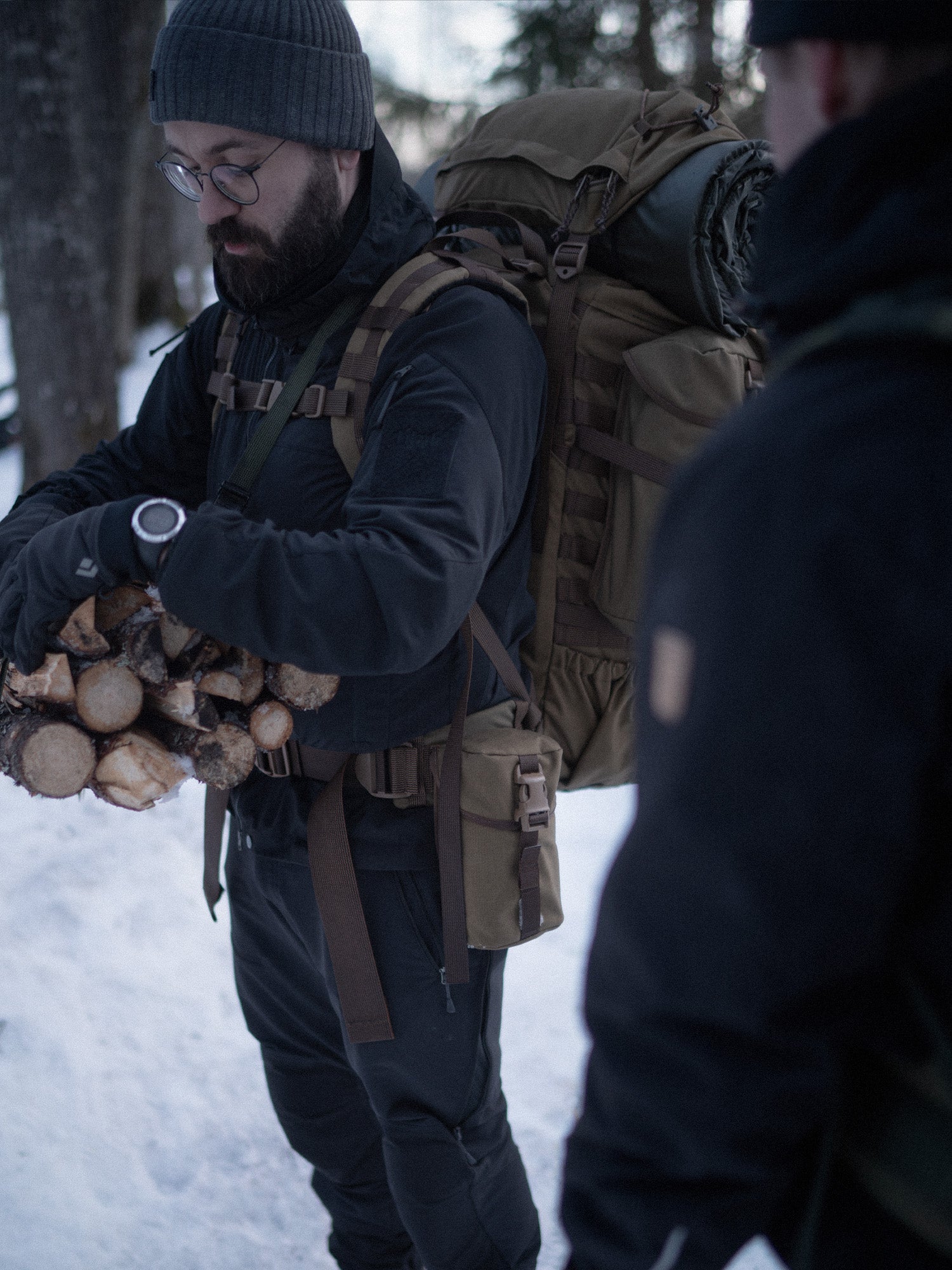 Person wearing a backpack with a Savotta Lidded GP pouch 3x4 attached, holding firewood in a snowy setting