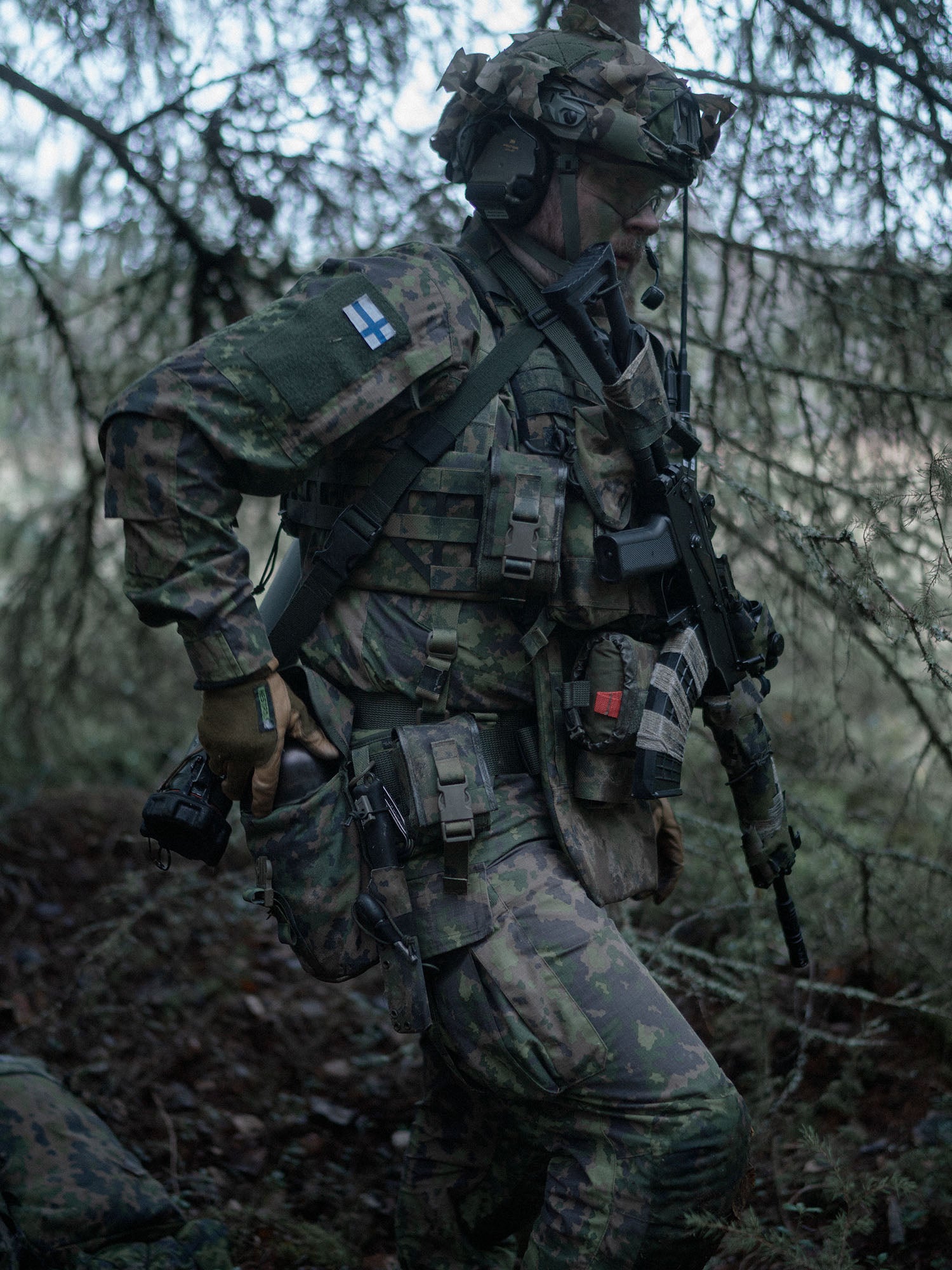 Soldier in camouflage gear with a rifle in a forest setting putting an item into a Savotta Lidded GP pouch 3x4.