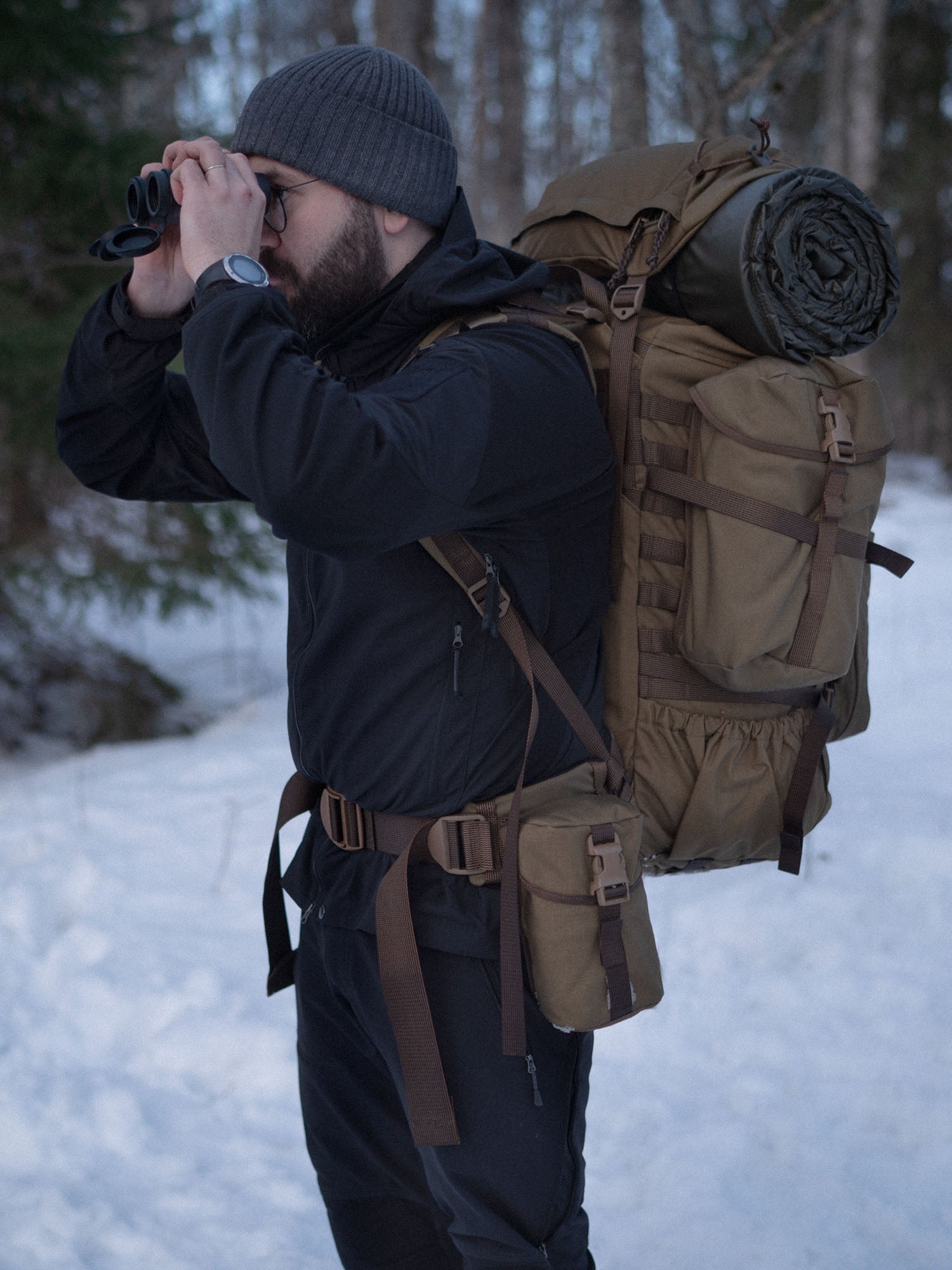 Man with binoculars and a Savotta Kantamus backpack with a Savotta Lidded GP pouch 4x5 attached to the side in a snowy forest