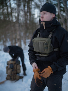 Person in outdoor gear standing in a snowy forest with another person in the background, wearing Savotta Rintamus chest pack in green.