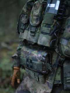 Close-up of a soldier's torso wearing a Savotta Varjelus plate carrier with Varjelus 3x2 AR15-AK placard and Varjelus groin flap attached, in a forest setting