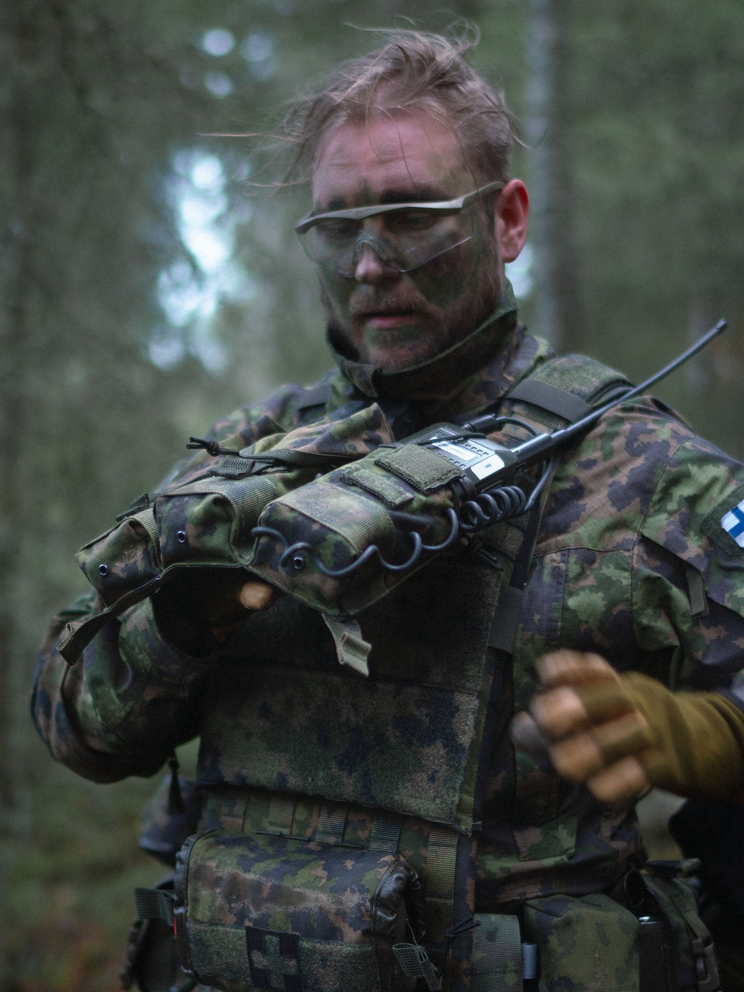 Person in military camouflage gear lifting the Savotta Varjelus 3x2 AR15-AK placard from a Varjelus plate carrier in a forest setting