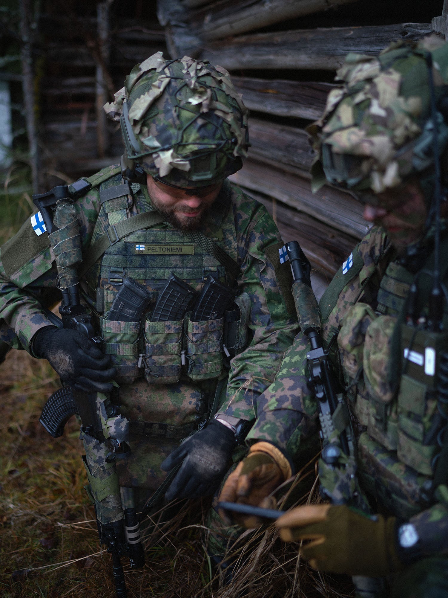 Two soldiers in camouflage gear with Savotta open top AR15-AK mag pouches attached, standing in front of a wooden cabin in a forest setting