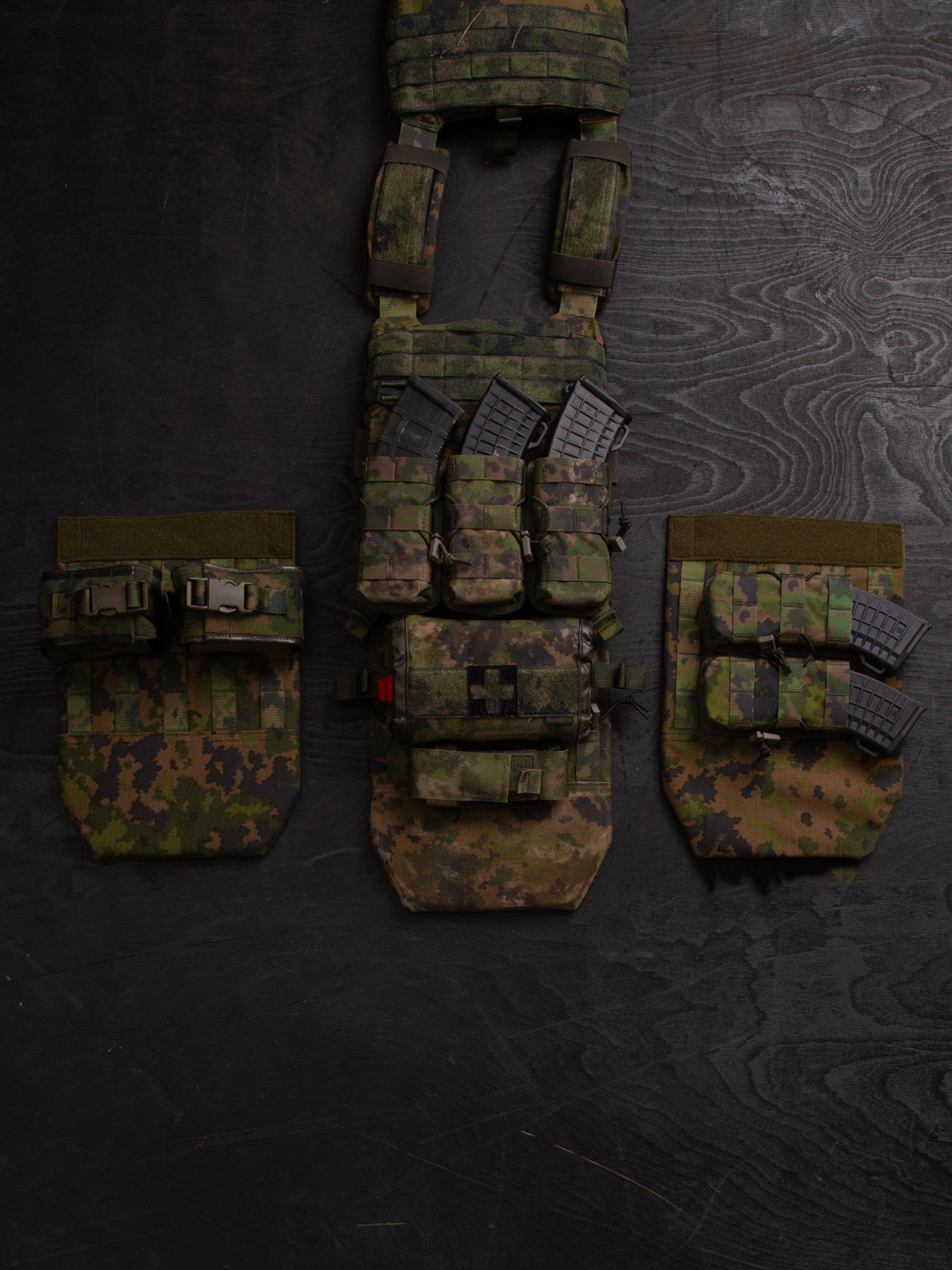 Three different setups you can do with a Savotta Varjelus groin flap. The Savotta plate carrier is being spread out on a dark wooden background. There are different Savotta mag pouches and Savotta S-IFAK horizontal mount attached to the groin flaps.