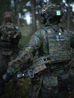 Person in camouflage military uniform adjusting the Savotta Varjelus plate carrier, in a forest setting