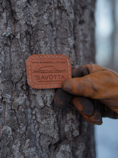 Hand wearing a brown glove holding a leather patch with 'Savotta' branding against a tree.