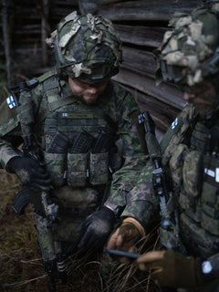 Two soldiers in camouflage gear with Savotta open top AR15-AK mag pouches attached, standing in front of a wooden cabin in a forest setting