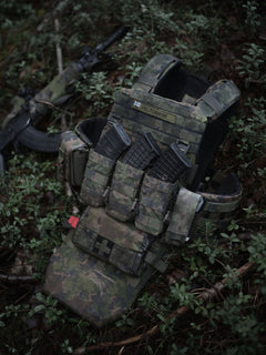 Camouflage tactical vest with Savotta open top AR15-AK mag pouches attached lying on a forest floor with a rifle in the background