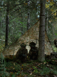 Two soldiers in camouflage gear sitting behind a Savotta Multicam Laavu tarp in a forest setting.