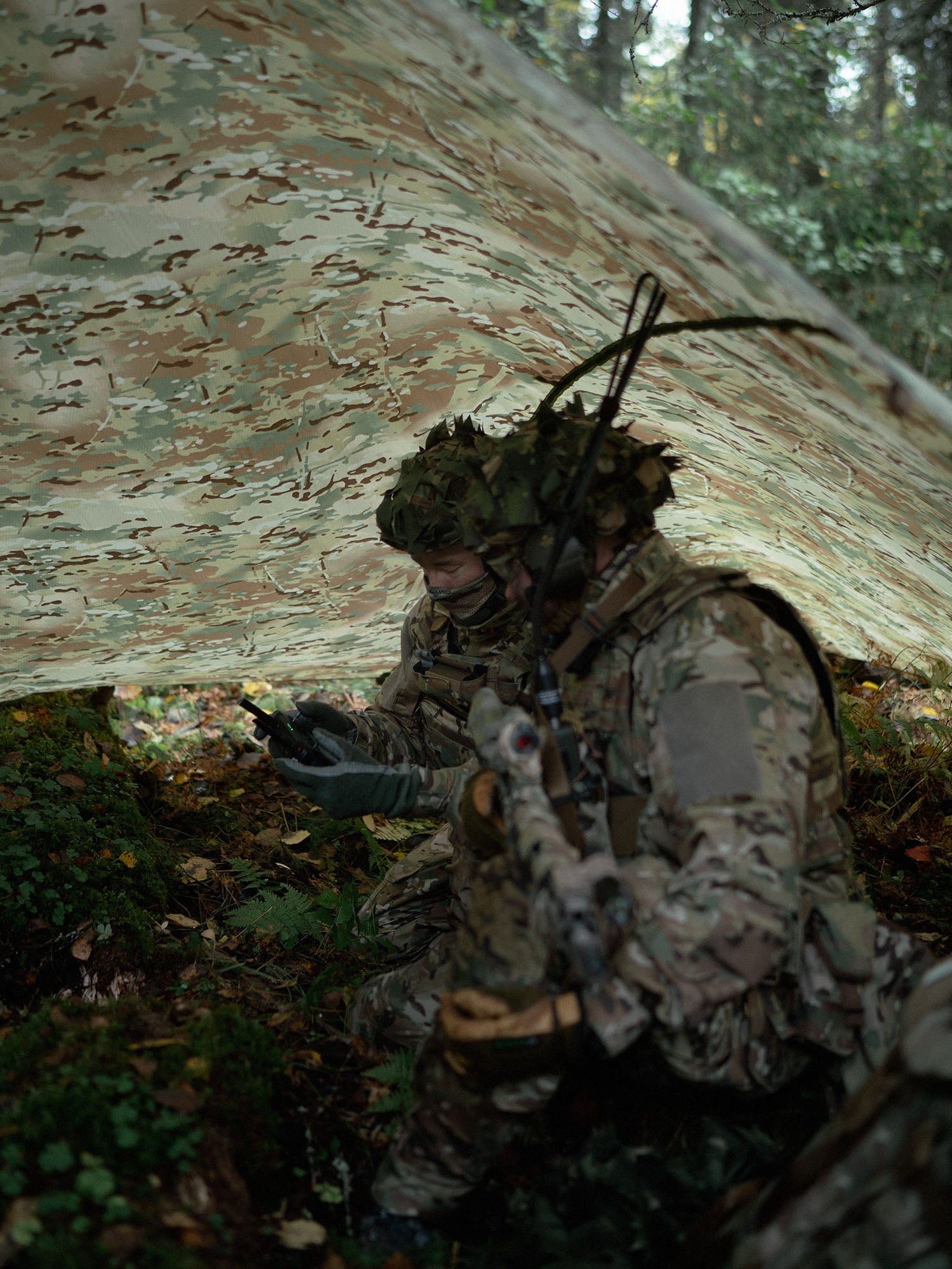 Two soldiers in camouflage gear sitting being a Savotta Multicam Laavu tarp in a forest setting.