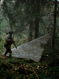 Person in camouflage gear walking by a Savotta Multicam Laavu tarp in a forest
