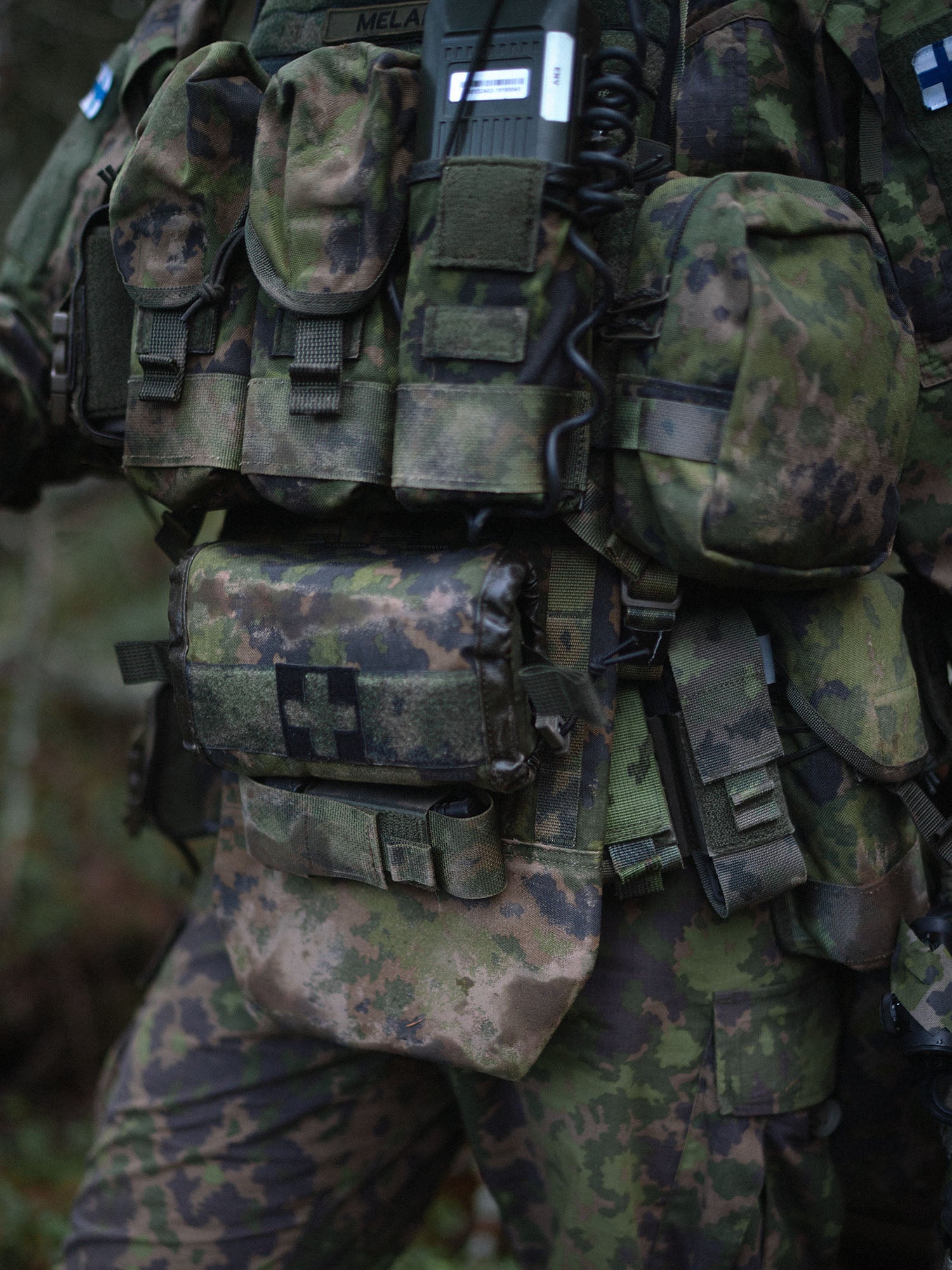 Person wearing camouflage gear with a Savotta Varjelus plate carrier with Varjelus groin flap and Varjelus 3x2 AR15-AK placard attached, in a forest setting