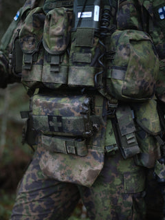 Person wearing camouflage gear with a Savotta Varjelus plate carrier with Varjelus groin flap and Varjelus 3x2 AR15-AK placard attached, in a forest setting