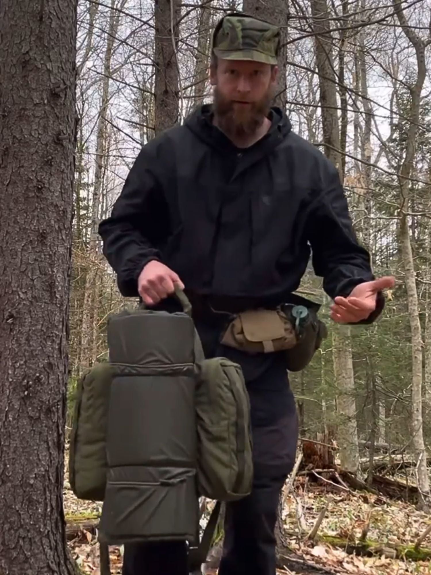 Man in outdoor clothing holding a backpack in a forest setting